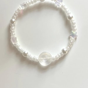 May include: A beaded bracelet featuring a circular arrangement of white pearl-like beads, translucent star-shaped beads, and a single clear, flat, circular disc bead positioned at the center. The beads are strung on a flexible cord in a circular loop.