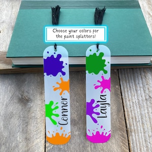 May include: Two personalized bookmarks with paint splatter designs. The bookmarks are white with colorful paint splatters and the names "Connor" and "Layla" written in black. The text "Choose your colors for the paint splatters!" is written on a white rectangle.