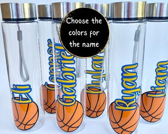 May include: Clear water bottles with silver lids and gray straps. Each bottle has a name in blue and yellow, along with an orange and black basketball graphic. The text on the image says, "Choose the colors for the name."