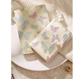 Può includere: Due quaderni color crema con un motivo a farfalla. Il quaderno più grande ha un design a farfalla blu chiaro e rosa, mentre il quaderno più piccolo ha un design a farfalla blu chiaro.