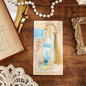 May include: A gold metal bookmark featuring a blue and white image of a sailboat and a tassel. The bookmark is displayed on a postcard with the text "Claude Monet" printed on it.