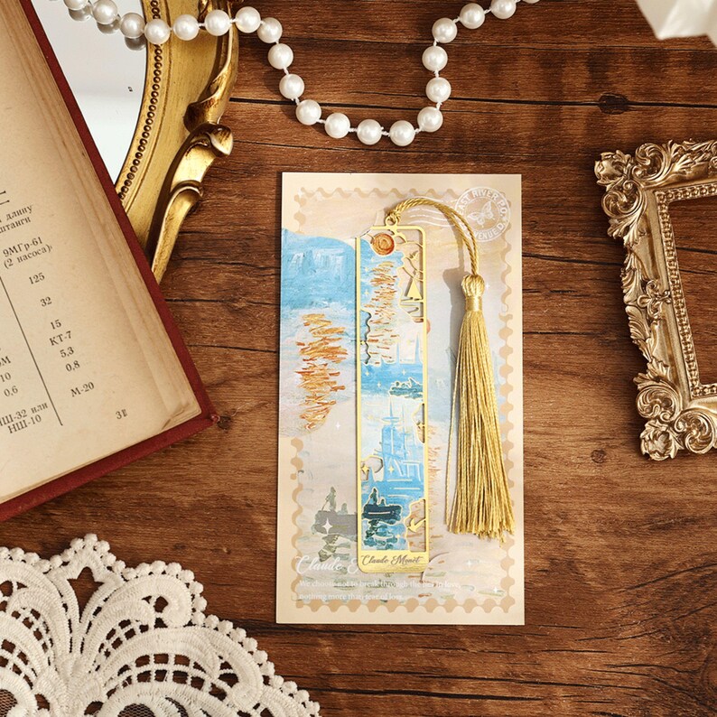May include: A gold metal bookmark featuring a blue and white image of a sailboat and a tassel. The bookmark is displayed on a postcard with the text "Claude Monet" printed on it.