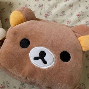 May include: A brown plush bear-shaped pouch with black eyes and a white and black nose. The bear has yellow ears and is made of soft, fuzzy material. The pouch is likely used for storing small items and has a zipper closure.