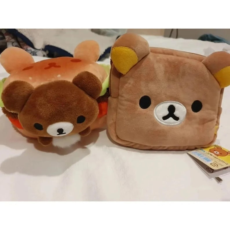 May include: Two plush toys: a brown bear-shaped burger and a brown bear-faced pouch. The burger has a light brown bun with red and orange details, and a dark brown bear face. The pouch is rectangular with a bear face and yellow ear accents.