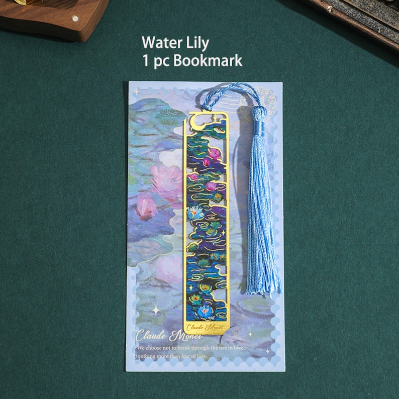 May include: A gold bookmark featuring a water lily design with blue and green tones. The bookmark has a blue tassel and the text "Water Lily 1 pc Bookmark" and "Claude Monet" is printed on the card.