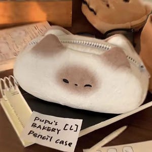 May include: A white and brown cat-shaped pencil case with a zip closure. The pencil case features a cute design with a brown spot and small black eyes. The text "Pupu's Bakery [CL] Pencil case" is on a small white paper.