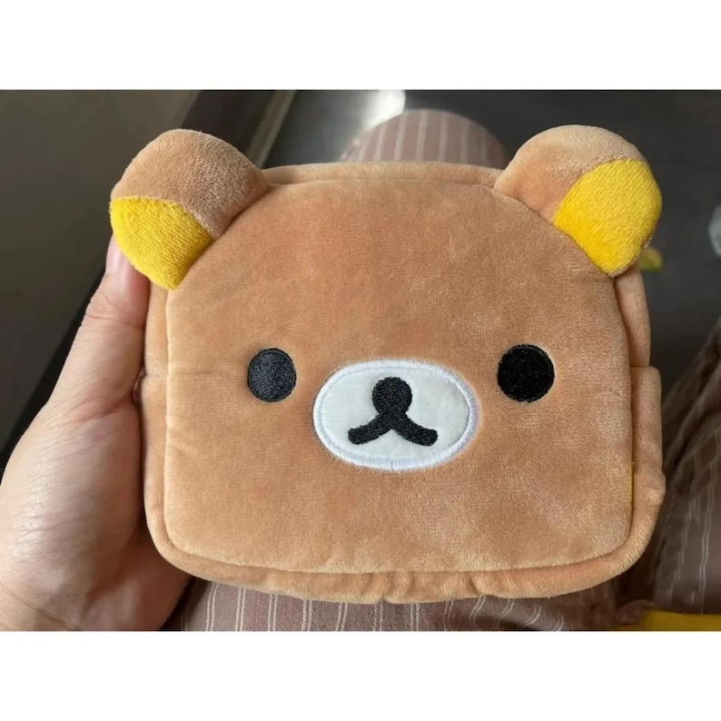 May include: A brown plush bear-shaped pouch with yellow ear accents. The pouch has black eyes and a white and black mouth design. The pouch is held by a hand and is likely a small accessory or storage item.