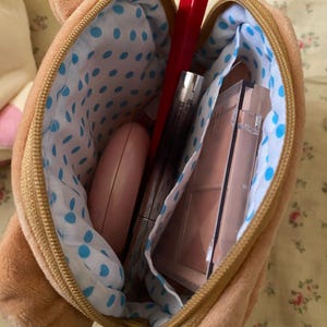 May include: A brown bear-shaped makeup bag with a gold zip, open to show various cosmetics. The interior lining is white with blue polka dots. Contents include red lipstick, mascara, and a compact.