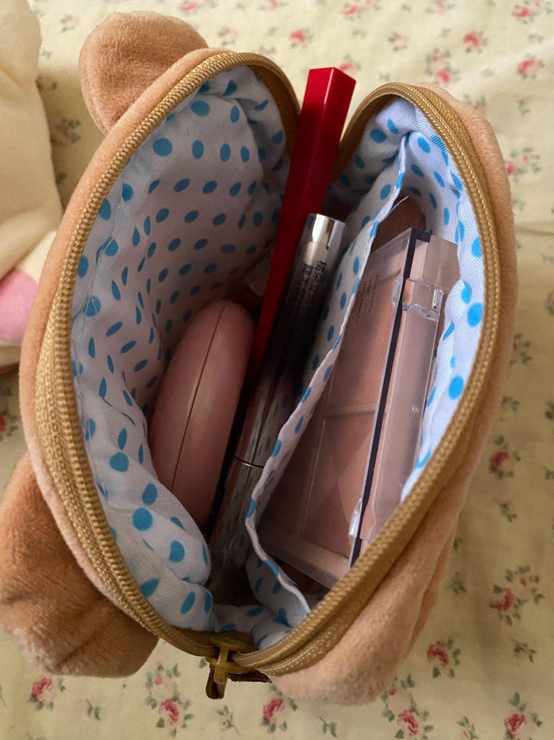 May include: A brown bear-shaped makeup bag with a gold zip, open to show various cosmetics. The interior lining is white with blue polka dots. Contents include red lipstick, mascara, and a compact.