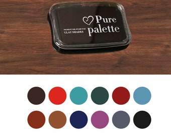 May include: A black ink pad with a clear plastic cover. The ink pad has the text "Pure Palette" and a heart symbol. The ink pad is on a wooden surface. The ink pad is surrounded by 12 color swatches.
