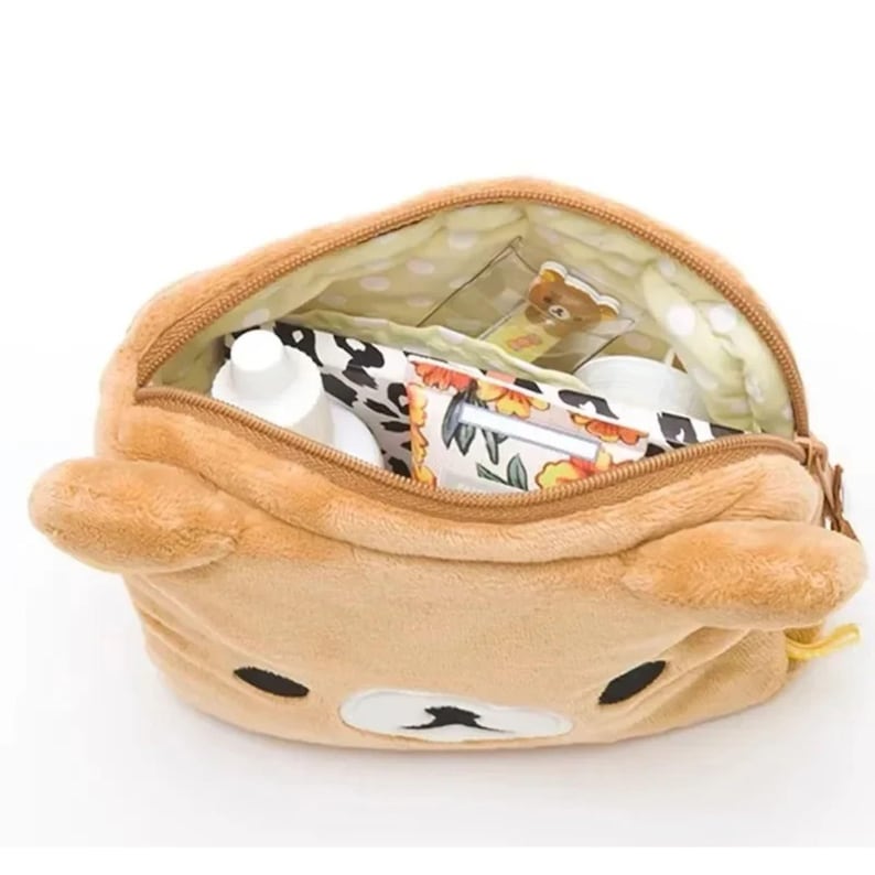 May include: A tan bear-shaped cosmetic bag with a zip closure. The bag is open, showing toiletries and a small bear-shaped tag. The interior lining is light yellow with white polka dots. The bag has black eyes and a white muzzle.