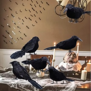 以下が含まれることがあります： ハロウィーンをテーマにしたディスプレイで、黒いカラスの装飾がいくつかあります。カラスは、ろうそく、頭蓋骨、クモの巣のような布が置かれたテーブルに配置されています。壁にはコウモリの切り抜きが飾られ、不気味な雰囲気を醸し出しています。