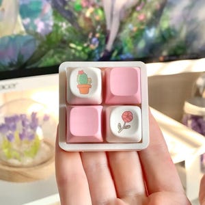 May include: Four pink and white keycaps with floral designs. The keycaps feature a cactus, a flower, and two blank keycaps.