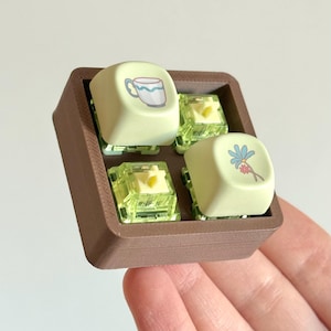 May include: A brown plastic tray holds four light green keycaps with different designs. The keycaps feature a cup of coffee, a flower, and two yellow squares.