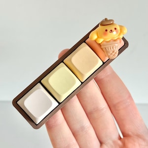 May include: A custom keyboard keycap set featuring four square keys in shades of white, yellow, and beige, set in a brown frame. A yellow cartoon dog with a brown hat and ice cream cone sits on the end. The keycaps are held in a hand.
