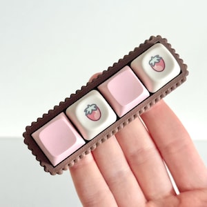 May include: A rectangular, brown keyboard keycap set with four keys. Two keys are white with a strawberry graphic, and two are solid pink. The keycaps are set in a brown, scalloped frame. The keycaps are a cute, decorative item.
