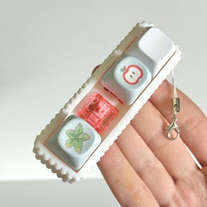 May include: A white rectangular tray with scalloped edges holds three keyboard keycaps and a pink switch. Two keycaps are light blue with an apple and leaf design. The third keycap is white. A silver clasp and string are attached.