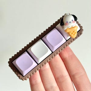 May include: A brown and white keycap set with a purple and white ice cream cone design. The keycaps are arranged in a row and feature a small, white dog with black ears on top of the ice cream cone.