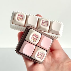 May include: Two sets of custom keyboard keycaps. One set is white with a scalloped edge, the other is brown with a scalloped edge. The keycaps feature pink and white designs, including a bow and a wrapped treat.