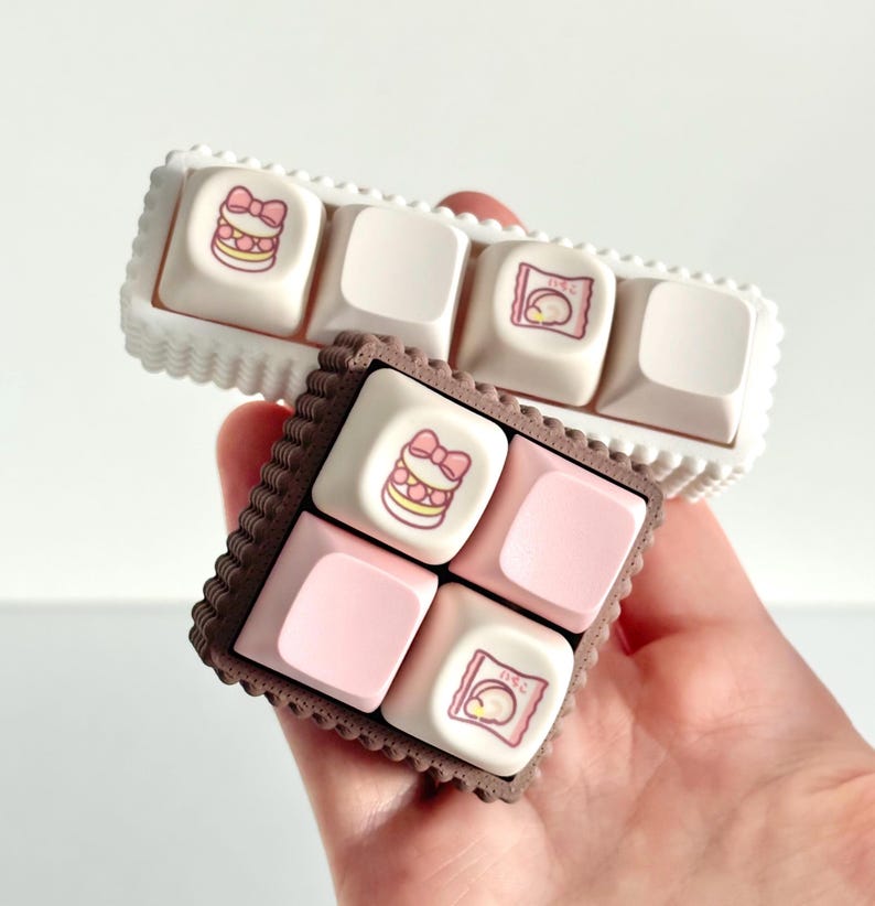May include: Two sets of custom keyboard keycaps. One set is white with a scalloped edge, the other is brown with a scalloped edge. The keycaps feature pink and white designs, including a bow and a wrapped treat.