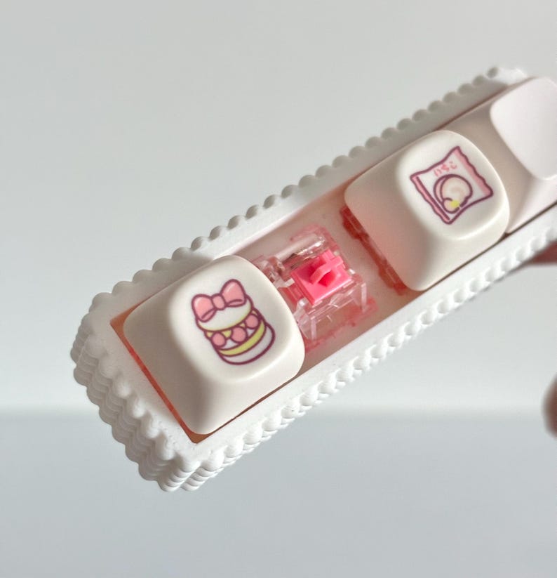 May include: A white keyboard key set with a pink and yellow macaron graphic and a pink bow. The keycaps are in a white tray with a scalloped edge. A pink switch is visible between the keycaps.