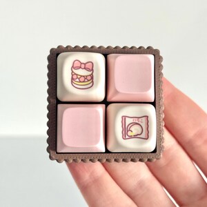 May include: A square, biscuit-shaped keyboard keycap set. The set features four individual keycaps, two pink and two white. One white keycap has a pink bow and cake design. The other white keycap has Japanese text.