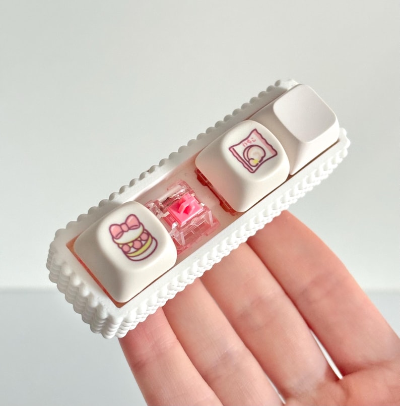 May include: A white keyboard key set with a decorative tray. The keys feature illustrations of a cake, a wrapped gift, and a blank key. A pink switch is visible between the keys. The tray has a scalloped edge.