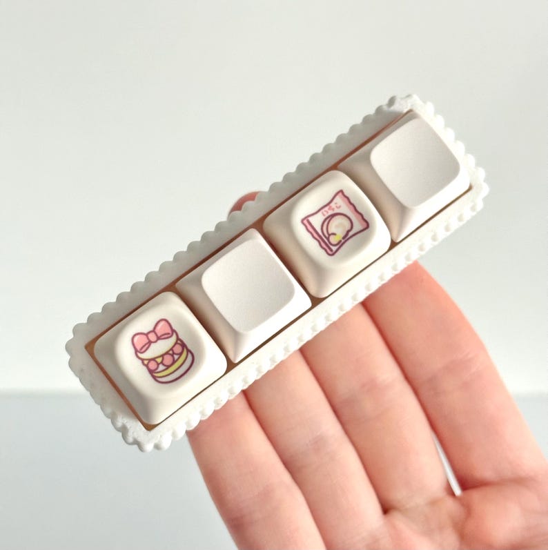 May include: A row of four off-white keyboard keycaps with decorative illustrations, set in a white scalloped frame. The keycaps feature illustrations of a bow, a wrapped item, and other designs in pink and yellow. The frame has a decorative edge.