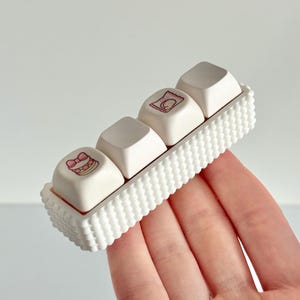 May include: Four white keyboard keycaps with small, colourful illustrations, held in a white, rectangular holder with a textured, scalloped edge. The illustrations include a cake and other food-related images. The holder is being held.