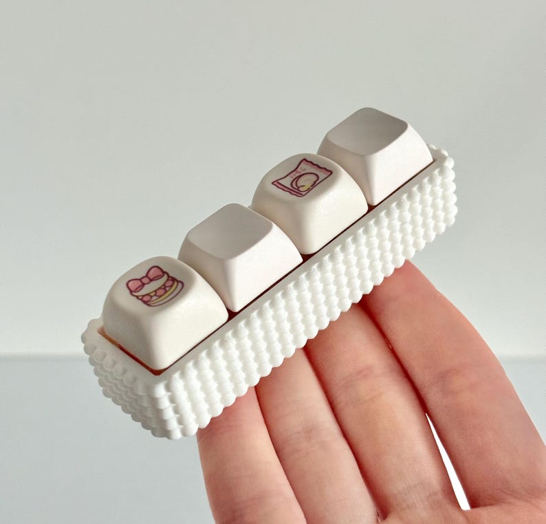 May include: Four white keyboard keycaps with small, colourful illustrations, held in a white, rectangular holder with a textured, scalloped edge. The illustrations include a cake and other food-related images. The holder is being held.