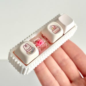 May include: A white plastic keycap holder with three keycaps featuring pink and white cake designs. The keycaps are in a row and are held in place by a white plastic frame.