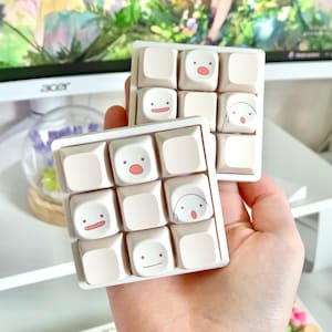 May include: Two sets of custom keyboard keycaps. Each set has nine light beige keycaps with various cartoon faces in pink and black. The keycaps are arranged in a 3x3 grid and are held in a white frame.