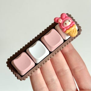 May include: A brown keycap set with four keycaps, two pink and two white. The keycaps are in a row and have a scalloped edge. A pink and white ice cream cone with a cartoon character on top is on the right side of the keycap set.