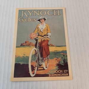 May include: A vintage advertisement for Kynoch Cycles featuring a woman in a yellow jacket and white hat riding a bicycle on a sandy path. The text reads "Kynoch Cycles" and "Kynoch Ltd Birmingham".