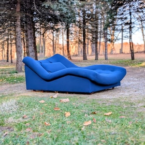 May include: A modern, blue chaise lounge with a curved design. The lounge is upholstered in a solid blue fabric and features a tufted seat. It is placed outdoors on a grassy area with trees in the background.