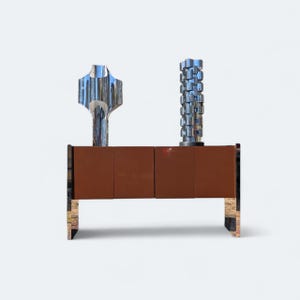 May include: A modern brown cabinet with four doors and chrome legs. Two chrome sculptures sit on top of the cabinet. One sculpture is a tall, vertical design, and the other is a more abstract shape.