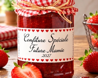 May include: A glass jar of red strawberry jam with a red and white checkered fabric lid cover tied with raffia. The label features red hearts and the text Confiture Spéciale Future Mamie 2027. Fresh strawberries and mint leaves surround the jar.