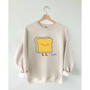May include: A beige sweatshirt with a cartoon illustration of a smiling slice of bread with legs and the word "PAN" printed below it.
