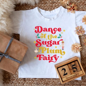 May include: White t-shirt with a red and gold graphic that says "Dance of the Sugar Plum Fairy". A wooden block calendar shows the date December 25th.