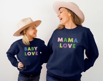 May include: A mother and daughter wearing matching navy blue sweatshirts. The mother's sweatshirt says "MAMA LOVE" in colorful letters. The daughter's sweatshirt says "BABY LOVE" in colorful letters. Both are wearing wide-brimmed hats.