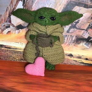 May include: A green crocheted Yoda doll holding a brown crocheted bowl. The doll is sitting on a brown surface with a pink crocheted heart in front of it.