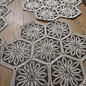 Peut inclure: Plusieurs dessous de plat hexagonaux avec un motif floral. Chaque dessous de plat présente un motif de fleur blanche en forme de marguerite sur un fond sombre. Les bords sont bordés d'une bordure décorative. Les dessous de plat sont disposés sur une surface en bois.