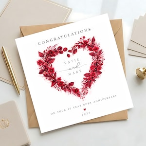 May include: A white anniversary card featuring a heart-shaped wreath of red flowers and foliage. The card is inscribed with "Congratulations", the names "Katie and Mark", and "On your 40 Year Ruby Anniversary", with the year 2026.