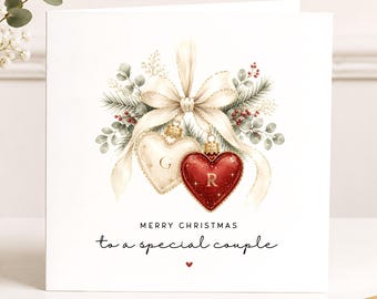 May include: A square white greeting card featuring a watercolor illustration of two heart-shaped ornaments, one cream with the letter G and one red with the letter R, tied with a cream ribbon, pine branches, and berries. Text reads MERRY CHRISTMAS to a special couple.