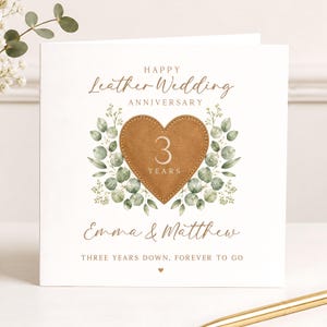 May include: A square white greeting card featuring the text Happy Leather Wedding Anniversary, a brown leather heart with the number 3 and the word YEARS, green eucalyptus leaf illustrations, the names Emma & Matthew, and Three years down, forever to go.