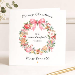 May include: A square white greeting card featuring a watercolour wreath of pine branches, a pink bow, a red apple, books, a gingerbread person, a mug, pencils, and stars. Text reads: Merry Christmas TO A wonderful TEACHER Miss Bennett.