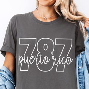 May include: Dark gray t-shirt with the text "787 puerto rico" in white. The shirt has short sleeves and a relaxed fit. A denim jacket is worn over the shirt. The shirt is a casual wear item.