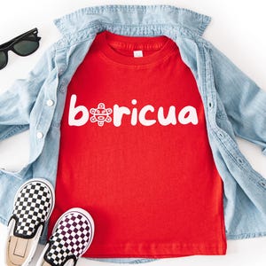 May include: A red t-shirt with the word "boricua" in white, along with a graphic. The shirt is layered under a light blue denim shirt. Black sunglasses and black and white checkered slip-on shoes complete the casual outfit.