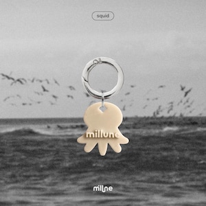 May include: A cream-colored squid-shaped keychain with the word "millune" in a serif font. The keychain is attached to a silver-colored metal ring. The word "squid" is in a white oval at the top. The background is a grayscale image of the ocean.