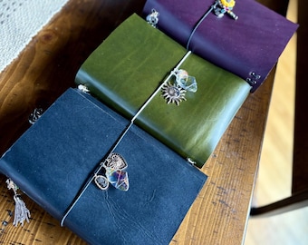 May include: A collection of leather journals in navy, olive green, and purple. Each journal is secured with an elastic band and adorned with decorative charms. The journals are arranged on a wooden surface.